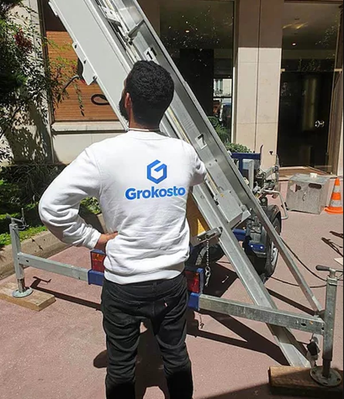 Déménageur Grokosto de dos, en sweat blanc au logo de la marque, devant un monte-meuble sur remorque installé au pied d’un immeuble