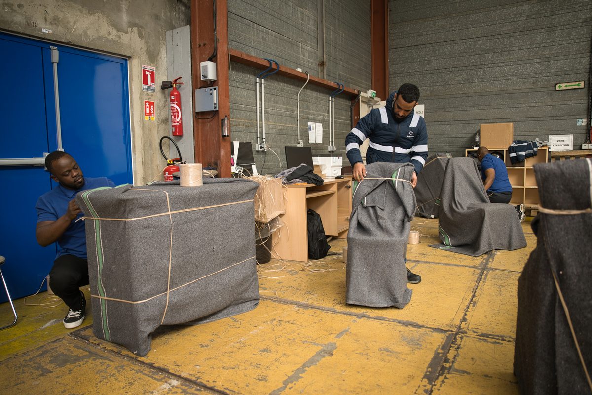 Déménageurs Grokosto enveloppant un meuble et des chaises dans des couvertures grises au sein d'un entrepôt