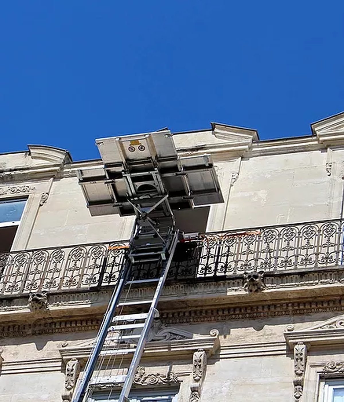 Monte-meuble dressé contre la façade en pierre d’un immeuble haussmannien, plateau arrivé au niveau d’un balcon en fer forgé